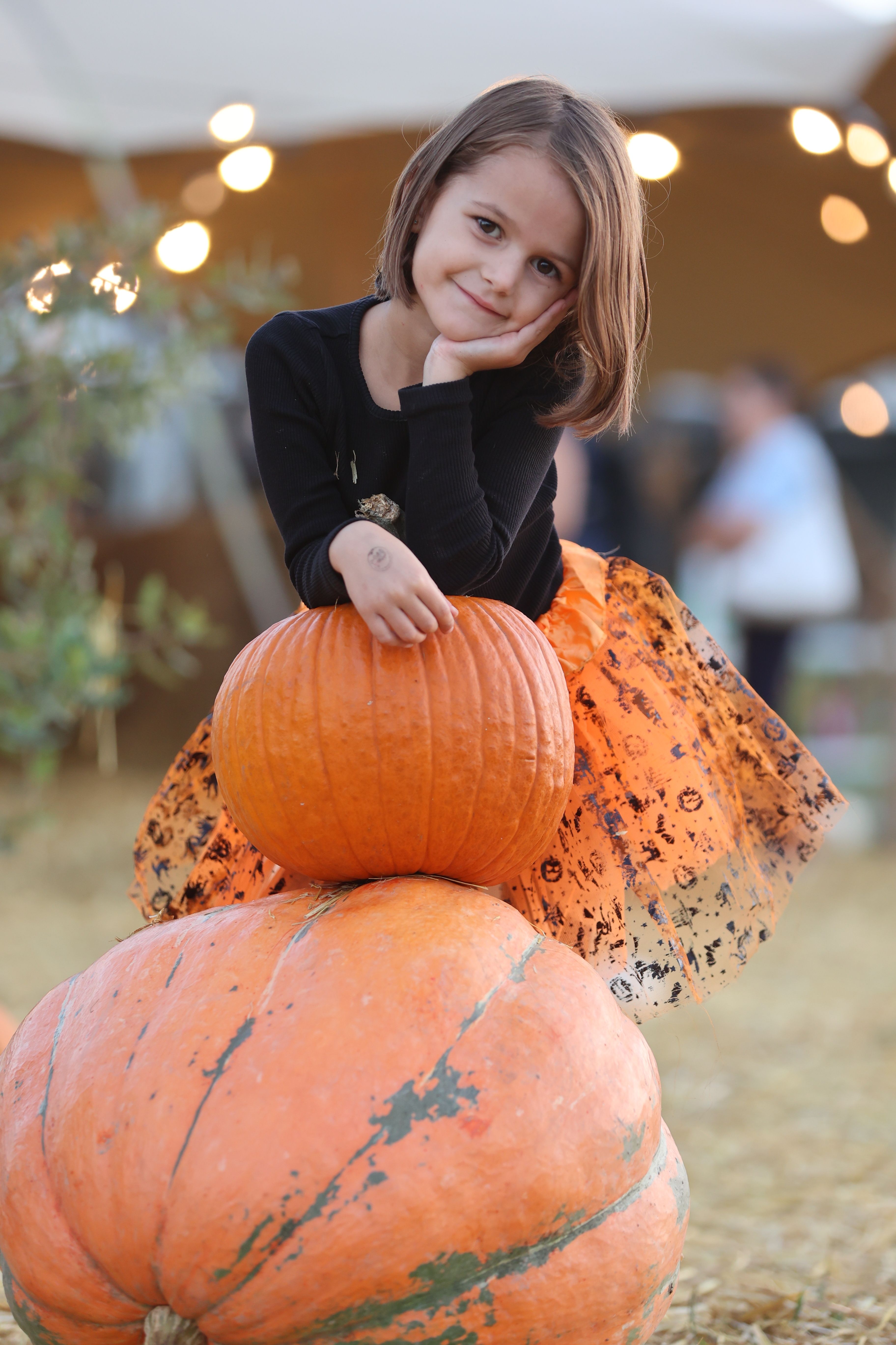 Young girl in a black top and orange tutu leaning on a stack of pumpkins at the Carlsbad Pumpkin Patch